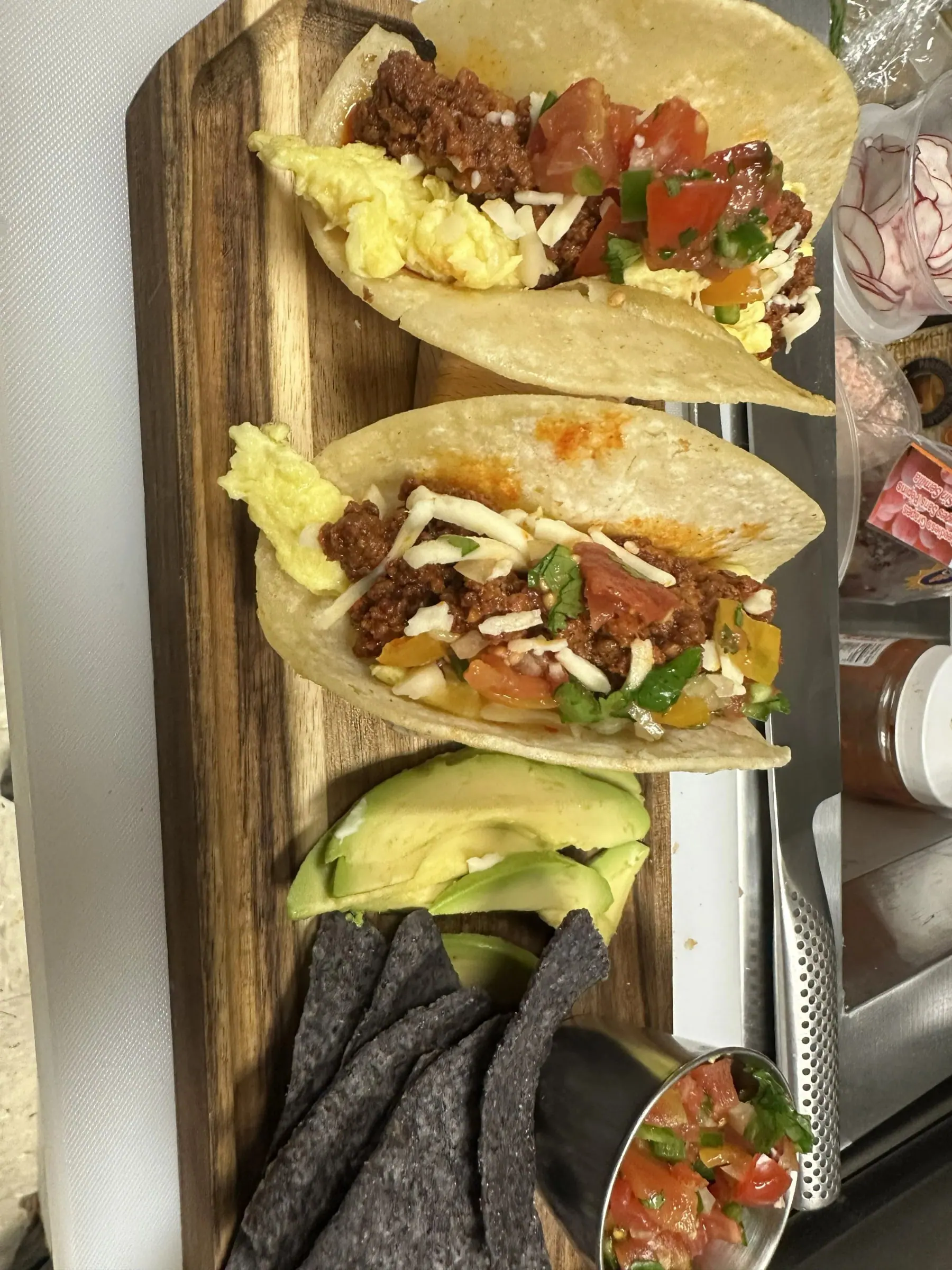 Soft tacos filled with chorizo, egg and pico de gallo on a wooden board with blue corn chips and avocado